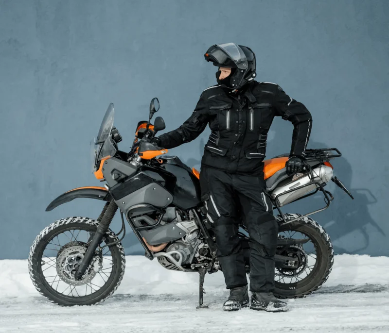 A man in black and orange gear stands beside a motorcycle, showcasing a sporty and adventurous look.